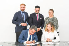 In the photo, five people are in a bright room at a glass table. In the foreground, one person in a navy blue suit is signing documents, and next to him sits a person in a white shirt looking at the documents. 