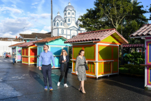 The photo shows three people walking down a cobbled alley surrounded by colorful wooden stalls with red roofs. In the background, there is a white church with several domes and trees. 