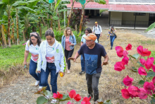 The photo shows a group of seven people walking uphill on a rocky path surrounded by vegetation. In the foreground, bright pink flowers are visible, and banana trees grow on the left. 