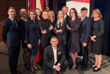 A group of people in elegant attire, standing in two rows against a backdrop of flags and a sign with the Harvard Medical School logo. 