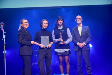Four people on stage. Two members of the HepaSwiych band are holding awards – one is holding a statuette, the other a framed diploma.