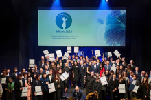A group of several dozen people stand on stage in elegant attire, holding diplomas and statuettes. In the background, there is a large screen with the inscription “StRuNa 2025” and information about the 15th edition of the National Student Scientific Movement Competition.