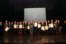 A group of elegantly dressed people. They are standing on stage and posing for a photo. Some of them are holding framed diplomas in their hands.