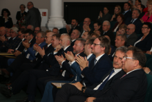 Participants at the gala. Elegantly dressed people sit in a large hall and applaud.
