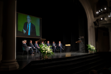 A group of elegantly dressed men are sitting on chairs on a stage in a large hall. They are talking. Behind them is a photo of Professor Zbigniew Religa.