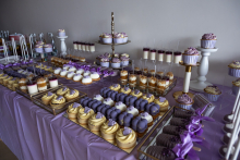 Stół jest przykryty fioletowym obrusem. Na nim stoją tace z małymi deserami w kolorach fioletowym, białym i złotym. 