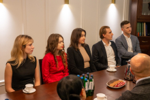 Five young people are sitting at a table in a conference room. There are cups, a jug, a plate of sweets, and bottles of water on the table.