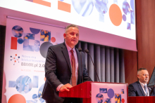 A man at the podium during a conference. Next to him is another man sitting behind a conference desk. In the background, there is a roll-up banner with information about the conference and a fragment of a multimedia screen.