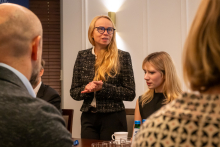 A person wearing an elegant jacket stands at a table in a conference room, hands folded in front of them.