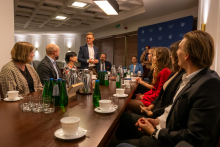 A person is standing at a table in a conference room, addressing a group of participants. There are cups, glasses, bottles of mineral water, and metal jugs on the table. 