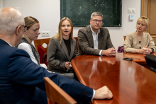 Grupa osób w czasie spotkania. Siedzą przy dużym brązowym stole. W centrum widać młodą kobietę, która coś opowiada.