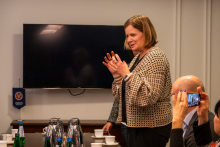 A person is standing at a table in a conference room, gesturing with their hands. There are cups, bottles of mineral water, and metal jugs on the table.