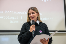 Młoda kobieta przemawia do mikrofonu. W ręce trzyma białą teczkę. Za nią widać ekran multimedialny.