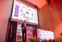 A man at the podium during a conference. Next to him is another man sitting behind a conference desk. In the background, there is a roll-up banner with information about the conference and a multimedia screen.
