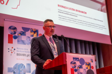A man at the podium during a conference. Next to him is another man sitting behind a conference desk. In the background, there is a roll-up banner with information about the conference and a fragment of a multimedia screen.