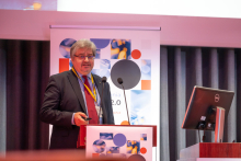 A man at the podium during a conference. Next to him is another man sitting behind a conference desk. In the background, there is a roll-up banner with information about the conference and a fragment of a multimedia screen.