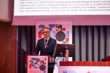 A man at the podium during a conference. Next to him is another man sitting behind a conference desk. In the background, there is a roll-up banner with information about the conference and a fragment of a multimedia screen.