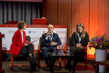 Several people sitting on chairs in an auditorium during a panel discussion. They are elegantly dressed. The man in the middle is holding a microphone and speaking.