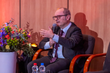 A man with a microphone in his right hand. His left hand is raised, gesturing. He is speaking.