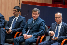 Trzech mężczyzn siedzi obok siebie na krzesłach w czasie panelu dyskusyjnego. Wszyscy są elegancko ubrani.