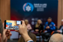 Osoba trzyma telefon i nagrywa panel dyskusyjny w sali konferencyjnej. Na ekranie telefonu widać kilka osób siedzących na krzesłach. W tle znajduje się granatowa ścianka z napisem ‘SAVE A LIFE – Światowy Zjazd Polonijnych Środowisk Medycznych.