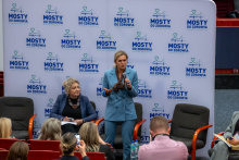 Dwie osoby uczestniczą w panelu dyskusyjnym na tle ścianki z napisem ‘Mosty do Zdrowia’. Jedna osoba siedzi i trzyma notatki, druga - Minister Edukacji stoi z mikrofonem i tabletem. Na pierwszym planie widać kilka osób w audytorium, część z nich robi zdjęcia. 