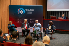 Cztery osoby siedzą na krzesłach w sali konferencyjnej podczas panelu dyskusyjnego. Jedna osoba trzyma mikrofon, pozostałe mają notatki. Uczestnicy noszą eleganckie stroje.  tle znajduje się granatowa ścianka z napisem ‘SAVE A LIFE – Światowy Zjazd Polonijnych Środowisk Medycznych’ oraz logotypami organizatorów. Na pierwszym planie widać kilka osób w audytorium.