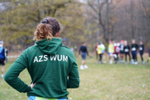 Kobieta ubrana w zieloną bluzę z napisem AZS WUM stoi odwrócona plecami do zdjęcia. W tle widać grupę ludzi. Wszyscy znajdują się w parku.