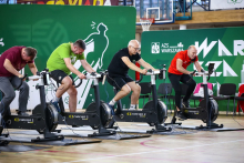 Grupa mężczyzn ubranych na sportowo, w tym rektor WUM, pedałuje na rowerkach stacjonarnych. W tle widać zielony baner z napisem AZS Warszawa.