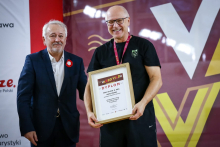 Dwóch mężczyzn, jeden ubrany elegancko, drugi - rektor WUM, ubrany na sportowo, pozują do zdjęcia. Rektor uśmiecha się szeroko, w rękach ma dyplom, na szyi medal. W tle widać baner z napisem Varsoviada.