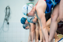 Grupa pływaczek na moment przed wskoczeniem do basenu. Mają na sobie czepki i okularki pływackie oraz kostiumy. Są pochylone do skoku.
