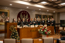 Grupa osób w eleganckich strojach wykonuje utwór chóralny w sali konferencyjnej ozdobionej świątecznymi dekoracjami. Na pierwszym planie stół z napisem „SALUT”, obok duża kompozycja kwiatowa i choinka. 