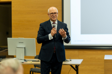 Prof. Rafał Krenke, elegancko ubrany, stoi na środku sali i przemawia. W prawej ręce ma mikrofon, za nim widać kawałek ekranu multimedialnego i komputer.