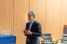 Elegancko ubrany mężczyzna stoi na środku auli i przemawia. W dali widać stół, na którym stoją kryształowe statuetki.