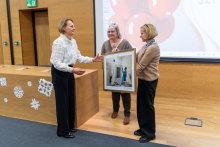 Trzy osoby stoją w sali z drewnianymi ścianami, obok dużego ekranu z prezentacją. Jedna osoba wręcza oprawione zdjęcie drugiej, a trzecia stoi obok.