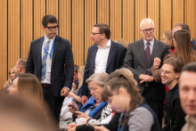 Grupa osób w sali konferencyjnej lub audytorium. W tle widoczne są pionowe drewniane panele na ścianie. W centralnej części kadru stoją trzy osoby ubrane w formalne stroje – garnitury z krawatami i koszulami.