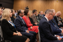 Osoby siedzące w rzędach na sali konferencyjnej. W tle znajdują się drewniane panele na ścianie, a w głębi kadru widoczny jest operator kamery. Uczestnicy mają na sobie eleganckie stroje – garnitury, marynarki oraz sukienki,
