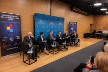 Panel dyskusyjny w sali konferencyjnej, w którym uczestniczy sześć osób siedzących w czarnych fotelach ustawionych w rzędzie. Każdy fotel ma przed sobą mały stolik z butelkami wody i szklankami. W tle znajduje się niebieska ścianka z napisami informującymi o jubileuszach.