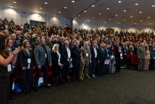 Bardzo duża grupa osób stojąca w dużej auli. Słuchają hymnu.