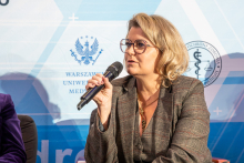 Elegancko ubrana kobieta siedzi na fotelu. W prawej dłoni trzyma mikrofon, do którego przemawia. W tle widać ściankę z logotypami.