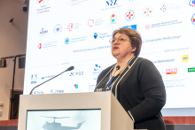 Elegancko ubrana kobieta za mównicą. Przemawia do mikrofonu. W tle widać ekran multimedialny z wieloma różnokolorowymi logotypami.