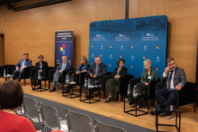 Osiem osób uczestniczących w panelu dyskusyjnym, zajmujących miejsca w czarnych fotelach ustawionych w rzędzie na drewnianej podłodze. Przed każdym fotelem znajduje się mały stolik z butelkami wody i szklankami. W tle widoczna jest niebieska ścianka z napisami informującymi o jubileuszach.