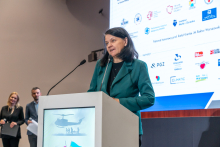 Elegancko ubrana kobieta stoi za mównicą i przemawia. W tle widać ekran multimedialny z różnymi logotypami.