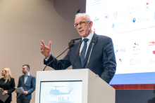 Elegancko ubrany mężczyzna stoi za mównicą i przemawia. Prawą rękę ma uniesioną do góry. W tle widać ekran multimedialny z różnymi logotypami.