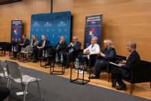 Panel dyskusyjny w sali konferencyjnej, w którym uczestniczy osiem osób siedzących w czarnych fotelach ustawionych w rzędzie. Przed każdym fotelem znajduje się mały stolik z butelkami wody i szklankami. W tle widoczna jest niebieska ścianka z napisami informującymi o jubileuszach.
