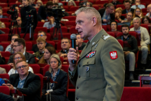 Mężczyzna ubrany w mundur z mikrofonem w ręku. W tle inne osoby, które siedzą na czerwonych krzesłach.