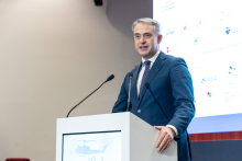 Elegancko ubrany mężczyzna stoi za mównicą i przemawia. W tle widać ekran multimedialny.