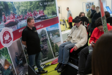 Stoisko promujące aktywności outdoorowe, m.in. rafting i paintball. Obok banera z informacjami o spływie pontonowym stoi mężczyzna, a inna osoba siedzi w fotelu z założonym zestawem VR. W tle widoczne są osoby obserwujące i korzystające z telefonów.