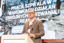 Elegancko ubrany mężczyzna stoi za mównicą i przemawia. W tle widać ekran multimedialny oraz wyświetlaną na nim prezentacją.