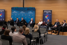 Panel dyskusyjny w sali konferencyjnej, w którym uczestniczy osiem osób siedzących w czarnych fotelach ustawionych w rzędzie. Przed każdym fotelem znajdują się małe stoliki z butelkami wody i szklankami. W tle widoczna jest niebieska ścianka z napisami informującymi o jubileuszach.